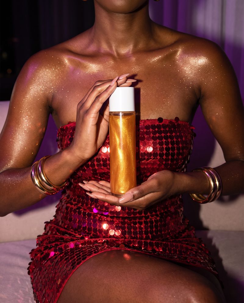 Gold shimmer on deep skin in a red sequin dress, holding a bottle of Molten Gold