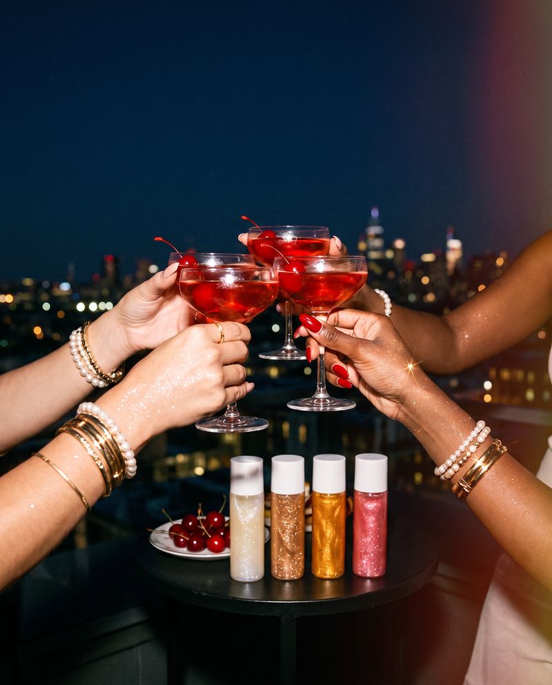 Three hands toasting with cherry cocktails on a rooftop at night, shimmer oils on the table